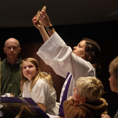 A female priest breaks the Communion bread while young people look up at it around the altar