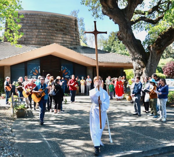 People gathered outside a church building, following a cross in procession